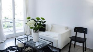 A modern living room with a white sofa, a glass coffee table, and a black chair. There is a window with white curtains and a view of greenery outside. - Best Located Executive Apartment in Maroussi
 (Marousi)