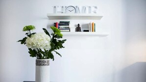 A white vase with white flowers on a glass table, a white shelf with books and a clock, and a word "HOME" on the wall. - Best Located Executive Apartment in Maroussi
 (Marousi)