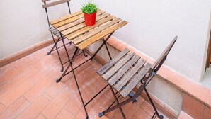 Une terrasse avec une table en bois et deux chaises, un pot de fleurs et une fenêtre avec des volets en bois.