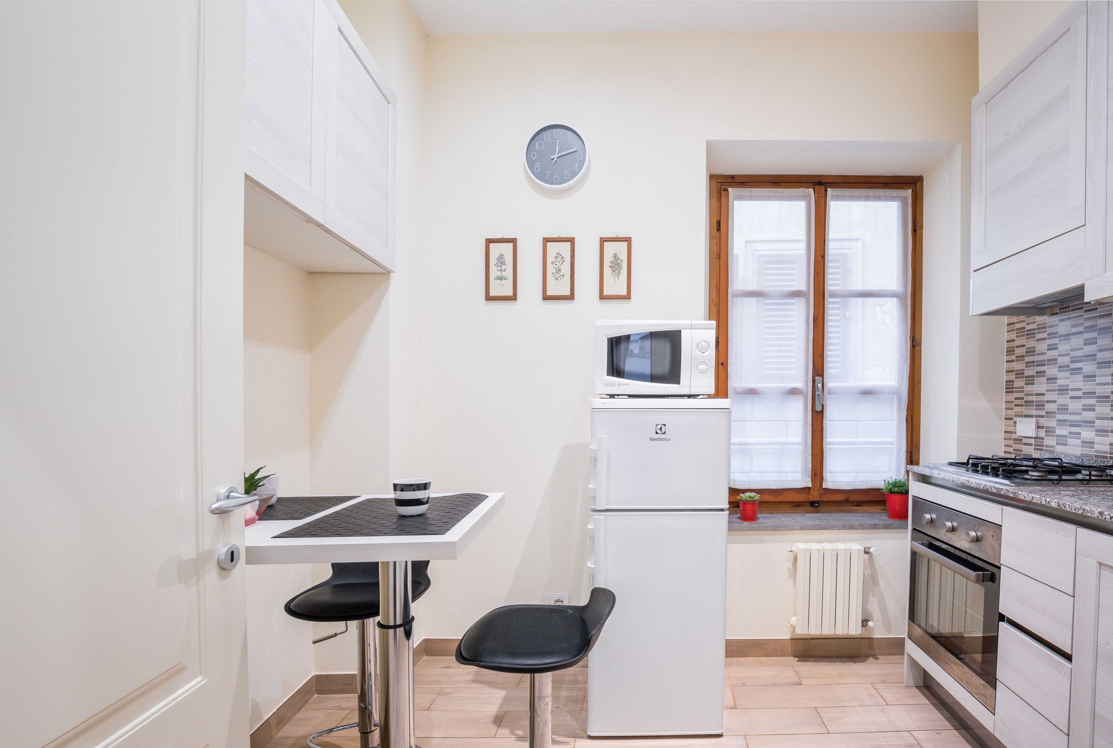 A compact kitchen with a microwave, refrigerator, and stove. There is a small dining table with a chair, and a clock on the wall.