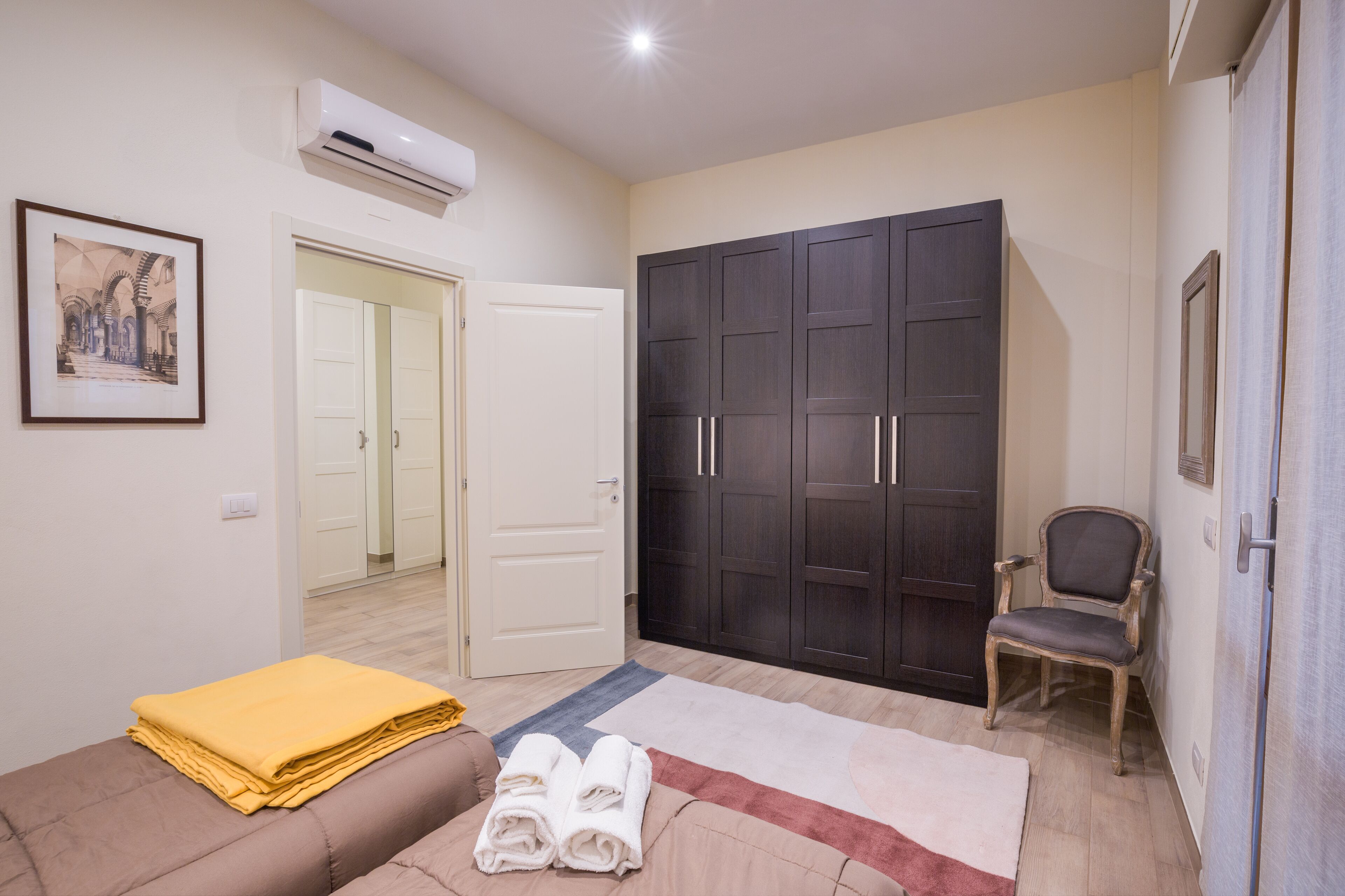 Une pièce comprenant un lit, une chaise, une armoire, un appareil de climatisation et un tableau encadré accroché au mur.