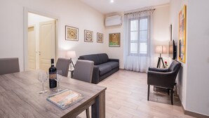 Un salon avec une table en bois, deux chaises, un canapé et une table basse. Les murs sont ornés d’œuvres d’art encadrées, il y a une lampe et une fenêtre avec des rideaux.