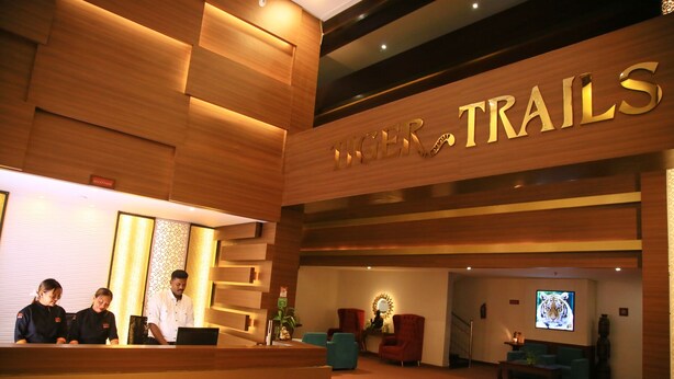 A reception area with a wooden reception desk, a sign reading "TRAILS", and a seating area with chairs and a television screen displaying a tiger. - Tiger Trails Thekkady (Peermade)