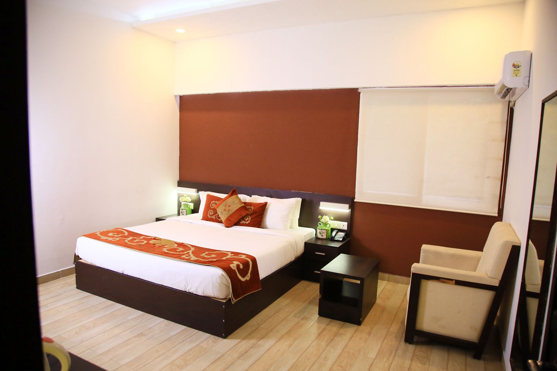 A hotel room with a bed, bedside table, chair, and a wall-mounted air conditioning unit.