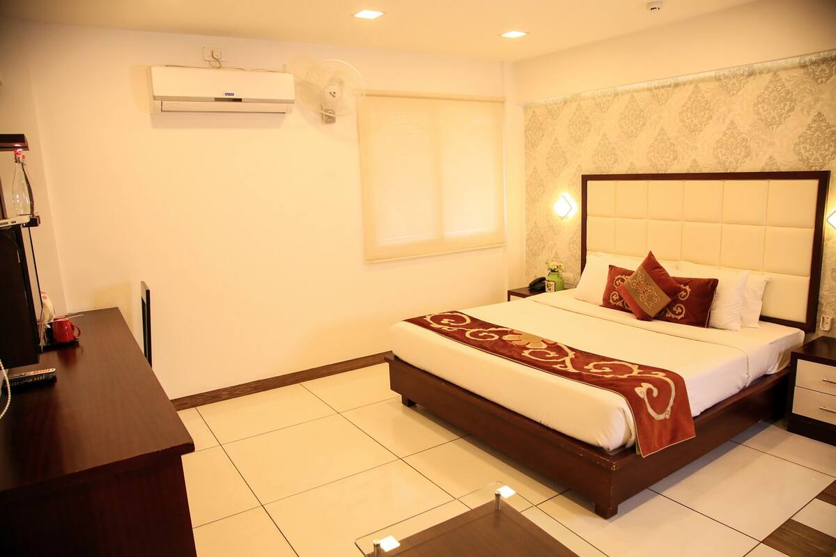 A hotel room with a large bed, a bedside table, a desk, and an air conditioning unit on the wall.