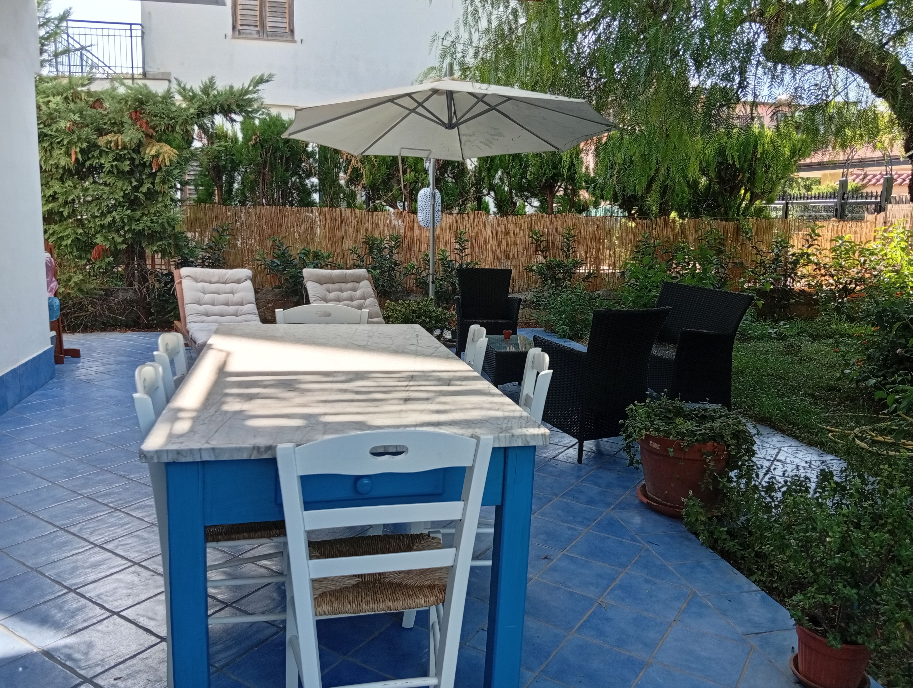 Un patio al aire libre con una mesa y sillas, un gran paraguas y diversas plantas y árboles.