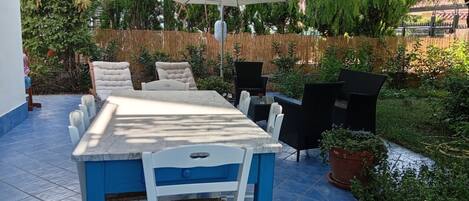 Un patio al aire libre con una mesa y sillas, un gran paraguas y diversas plantas y árboles.