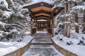 Une passerelle couverte en bois mène à un bâtiment doté d’une entrée en verre, le tout étant entouré d’arbres recouverts de neige.