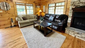 Interior acogedor de una cabaña con piso de madera, chimenea de piedra, sofá de cuero, sofá color beige y una mesa de centro.