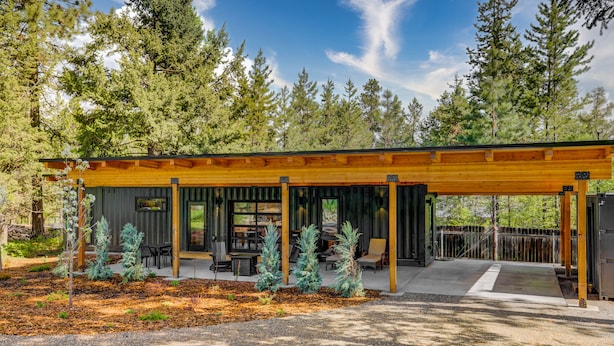 A modern wooden structure with a covered patio, outdoor seating, and a view of a forested area. - Two Pines - Unique container home w/hot tub - Downtown - Trailer Parking - Pet friendly (McCall)