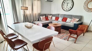 A modern living room with a sofa, a dining table with chairs, a coffee table, and a clock on the wall. - Superbe Appartement à Saint François Direct sur la Plage (Saint François)