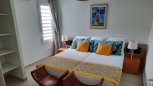 A neatly arranged bedroom with a bed, two chairs, a nightstand with lamps, a window with curtains, and a wall-mounted picture. - Superbe Appartement à Saint François Direct sur la Plage (Saint François)