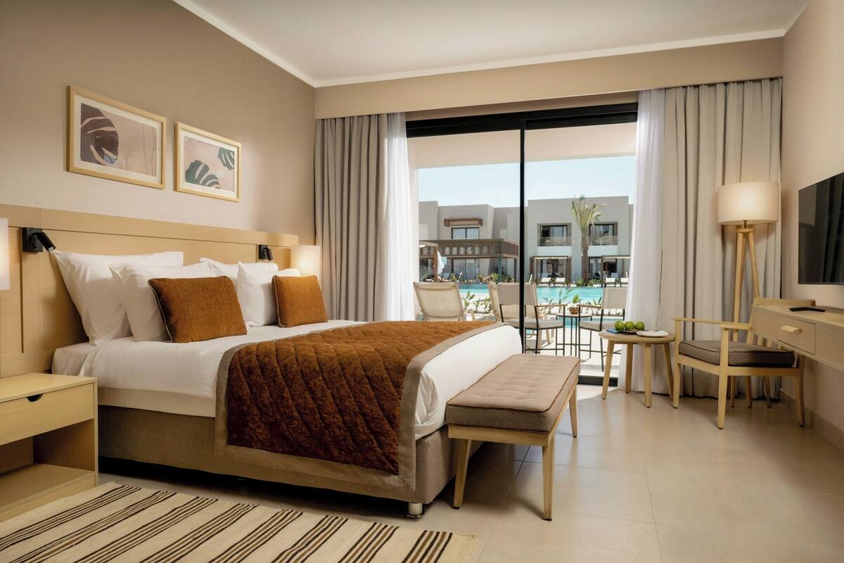 A hotel room with a bed, bedside tables, a desk, a chair, a TV, and a view of a pool and buildings.