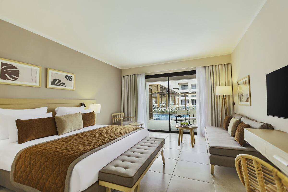 A hotel room with a large bed, a sofa, a small table, and a TV. There is a balcony with a view of buildings and a pool.