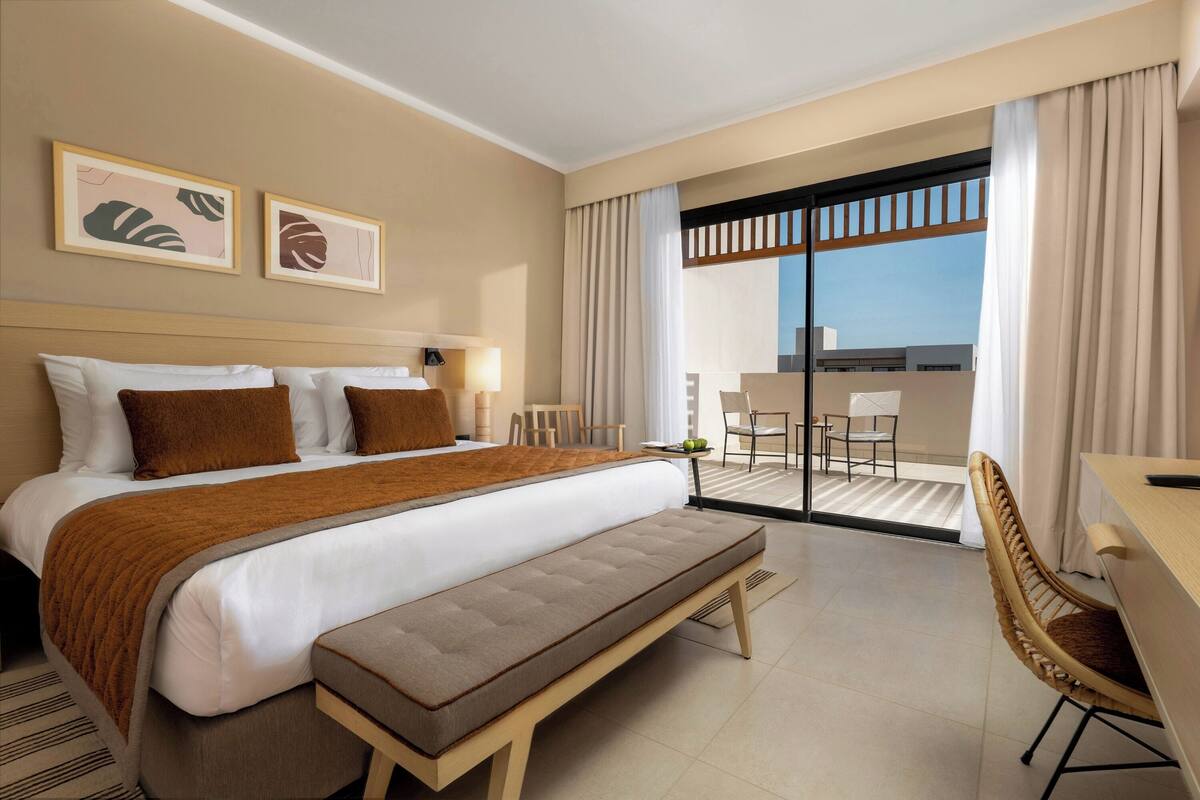 A hotel room with a large bed, a bench, a desk, and a balcony with a view.