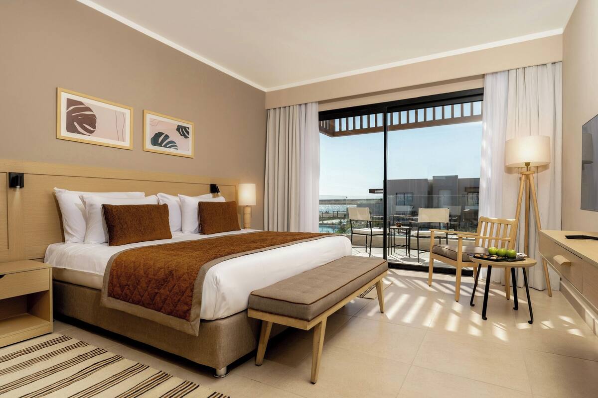 A hotel room with a large bed, a desk with a chair, a bench, and a view of the city through large windows.