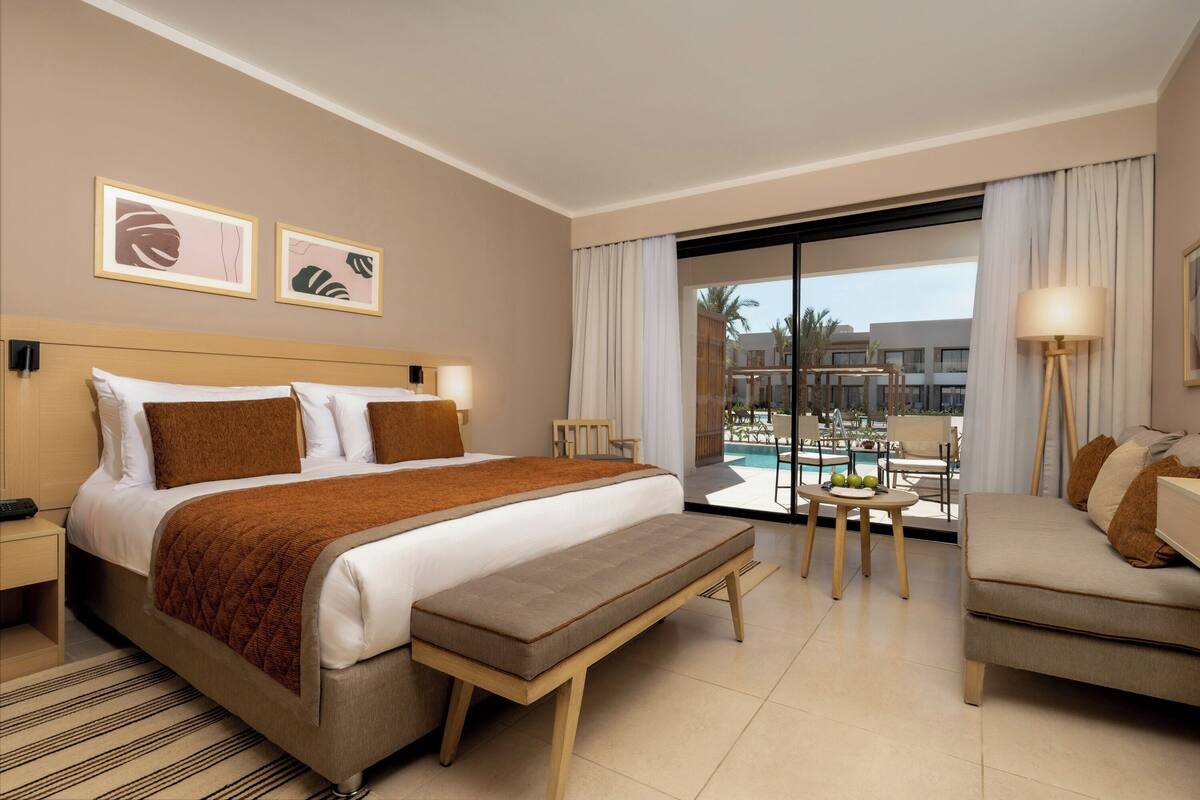 A hotel room with a large bed, a bench, a sofa, a dining table with chairs, and a view of a pool and buildings.