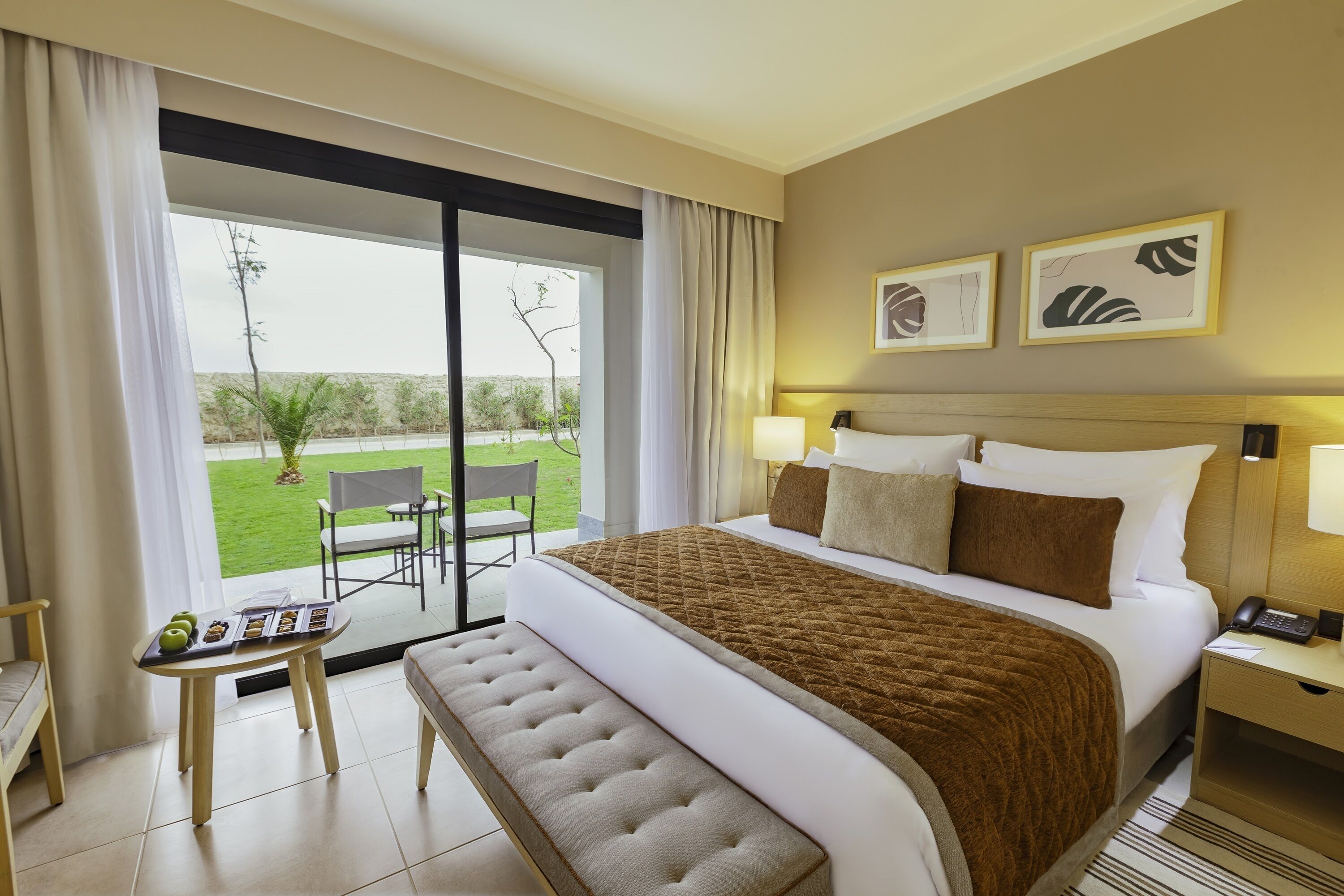A hotel room with a large bed, a bench, a small table with food, a chair, and a view of the outdoors.