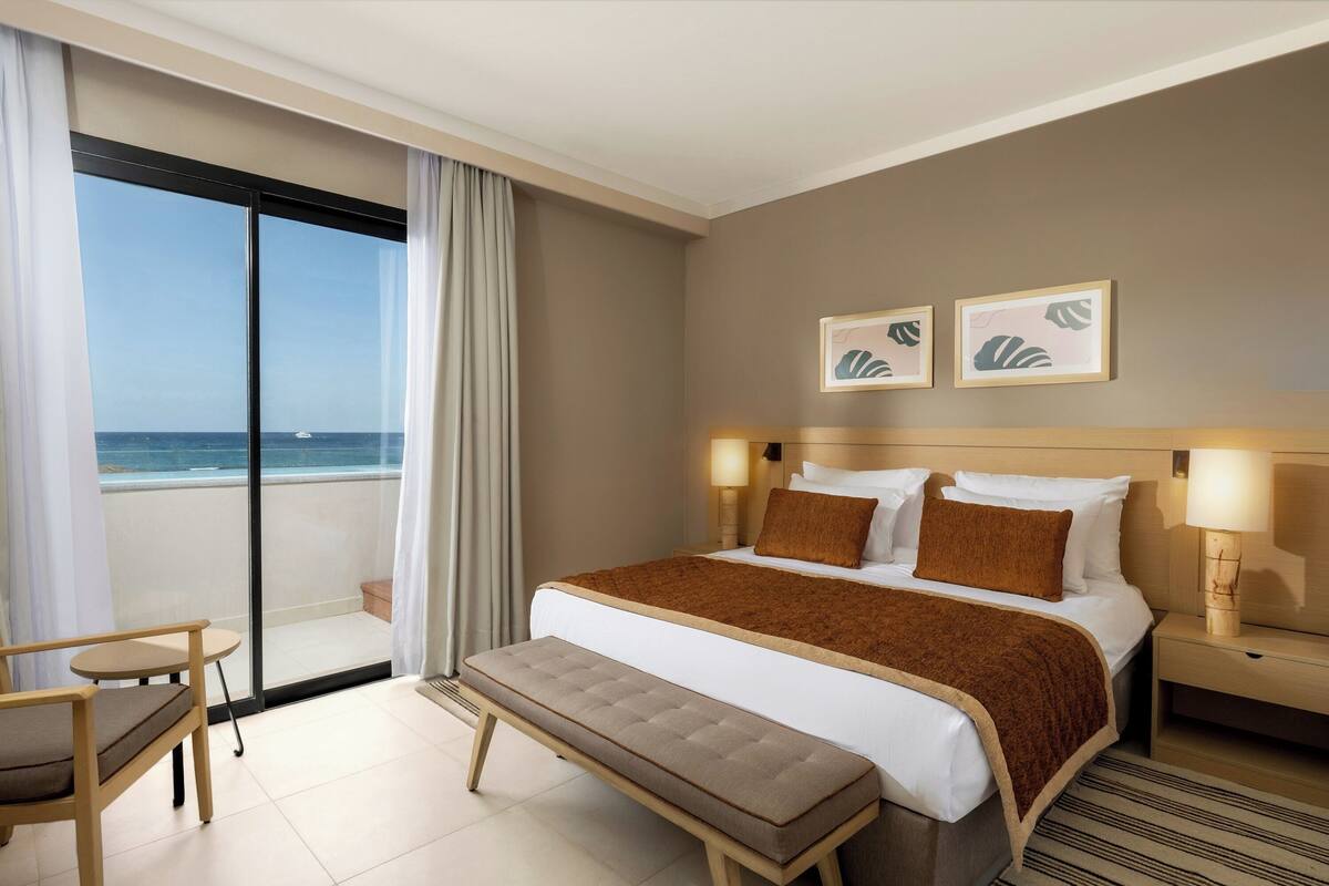A hotel room with a large bed, a bench, a chair, a nightstand, and a view of the ocean.