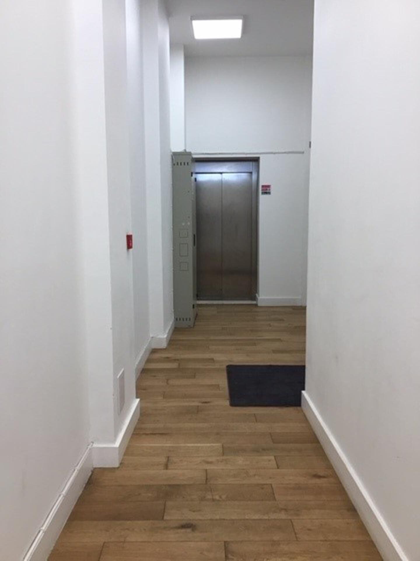 A narrow hallway with wooden flooring, white walls, and an elevator at the end.