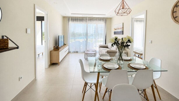 A modern dining area with a glass table, white chairs, and a central vase with flowers. - OCEAN HOMES - Apartamentos exclusivos en Isla Canela (Isla Canela)