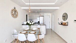 A modern kitchen with a central island, white cabinetry, and a dining area with a glass table and white chairs. - OCEAN HOMES - Apartamentos exclusivos en Isla Canela (Isla Canela)