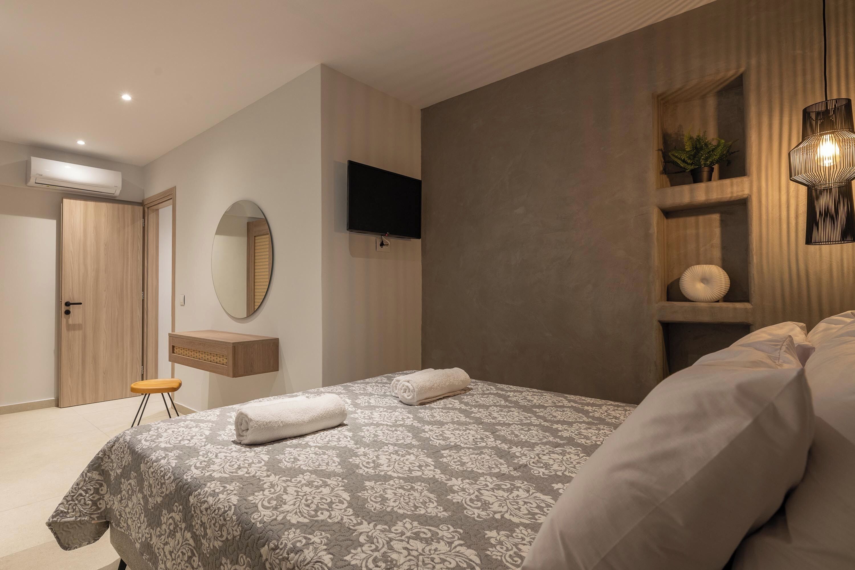A modern bedroom with a large bed, a TV mounted on the wall, a wooden wardrobe, and a small stool.