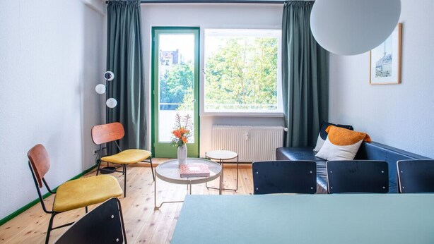 Quarto moderno com cama, sofá, mesa de jantar e cadeiras. Possui janela com cortinas, radiador e luminária pendente.