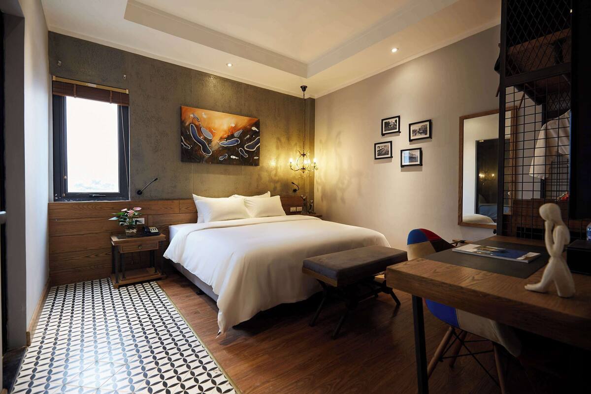 A hotel room with a large bed, a desk, a chair, a small table, a window, and artwork on the wall.