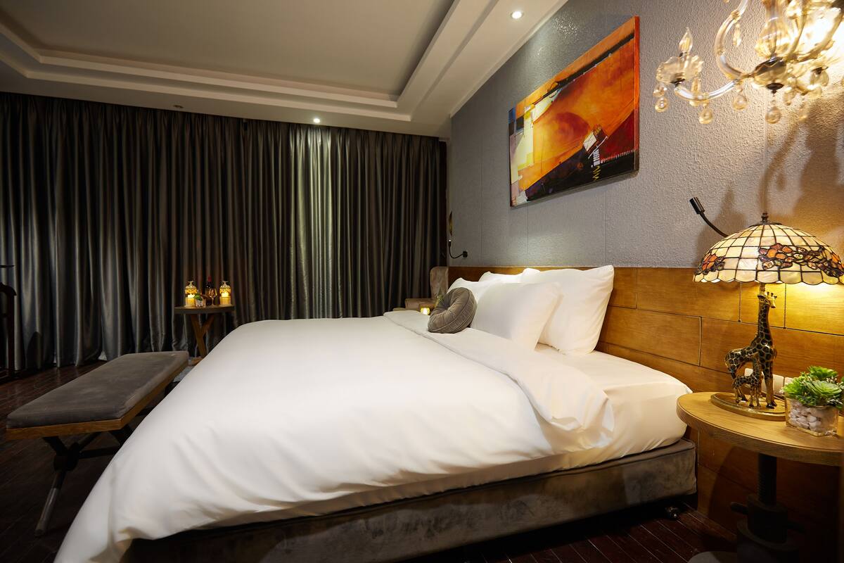 A hotel room with a large bed, a bench, a nightstand with a lamp, and a painting on the wall.