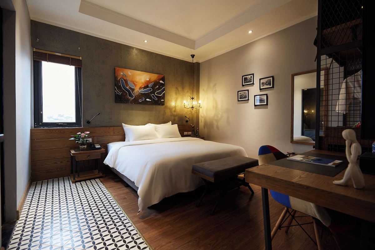 Una habitación de hotel con cama, mesita de noche, lámpara, obras de arte, cuadros enmarcados y un escritorio con silla.