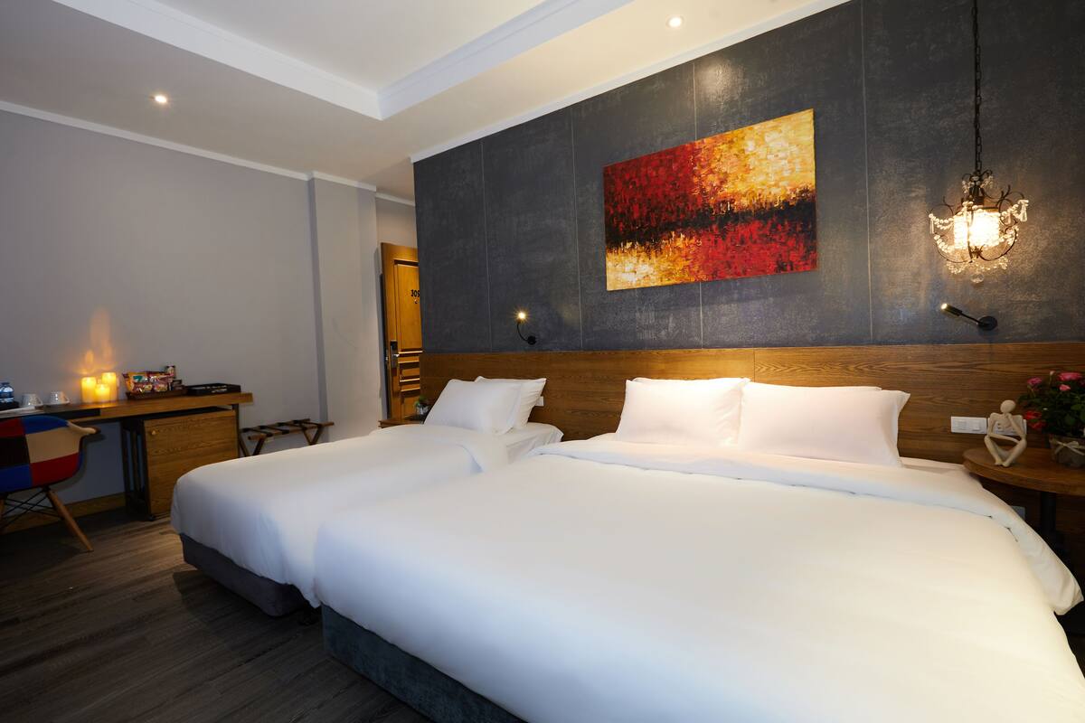 A hotel room with a large bed, a desk with a chair, a painting on the wall, and a chandelier.