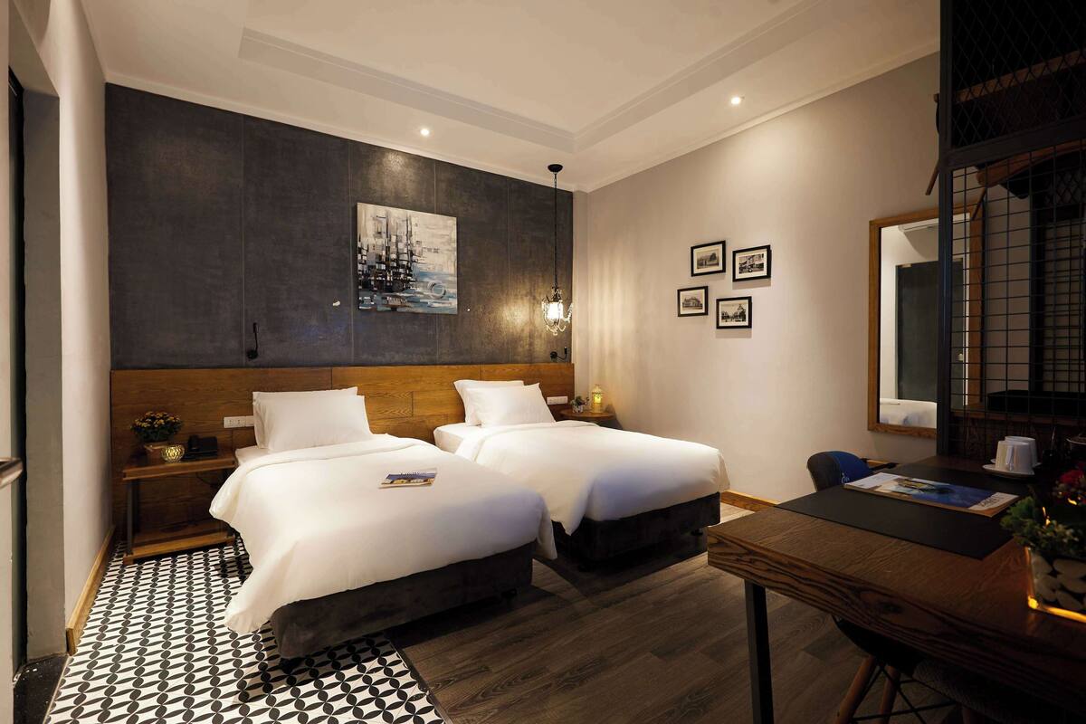 A hotel room with two beds, a wooden headboard, a black and white patterned rug, a desk with a chair, and a wall-mounted painting.
