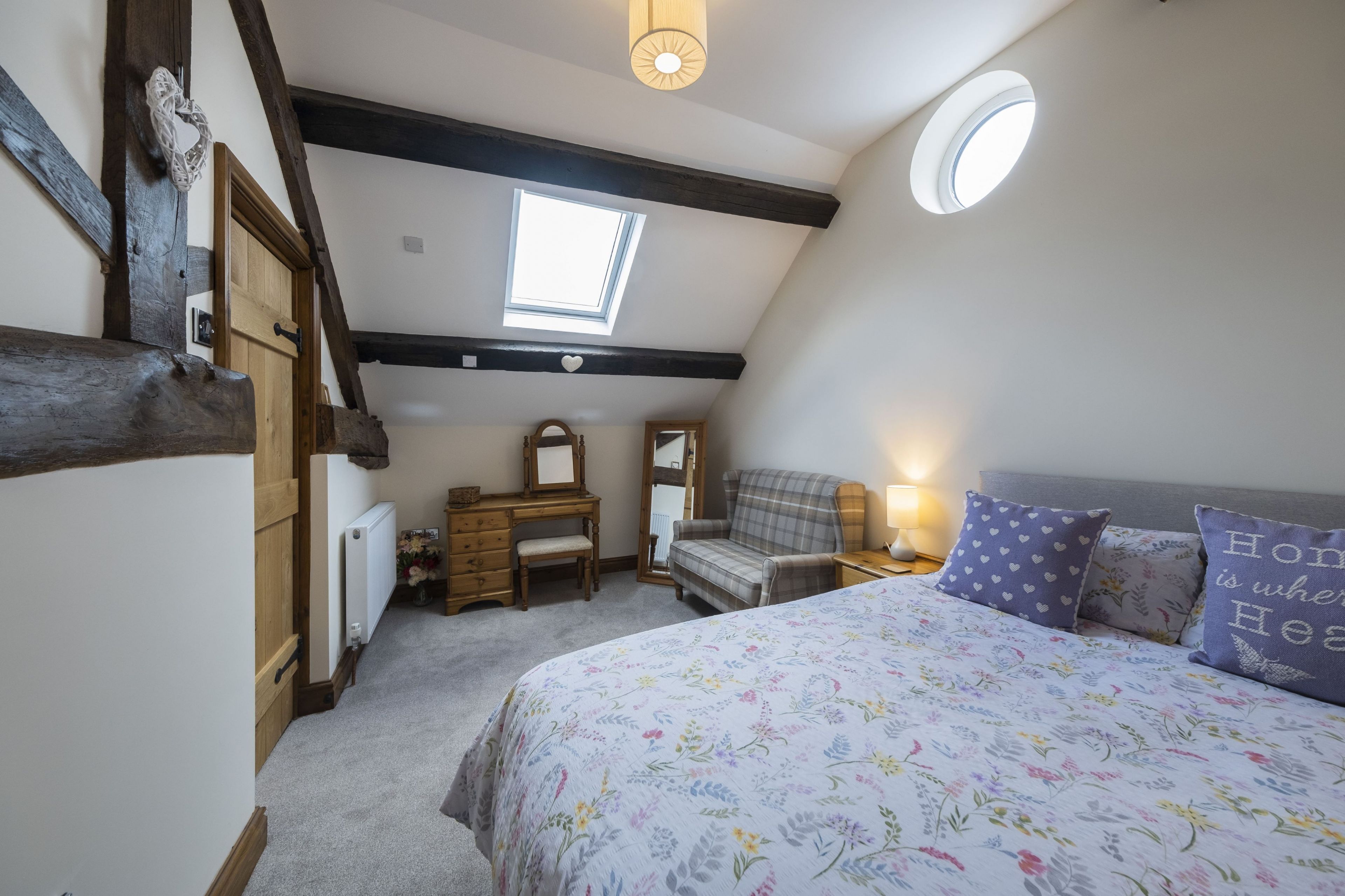 A bedroom with a sloped ceiling, a skylight, a bed with floral bedding, a dresser, a chair, and a mirror.
