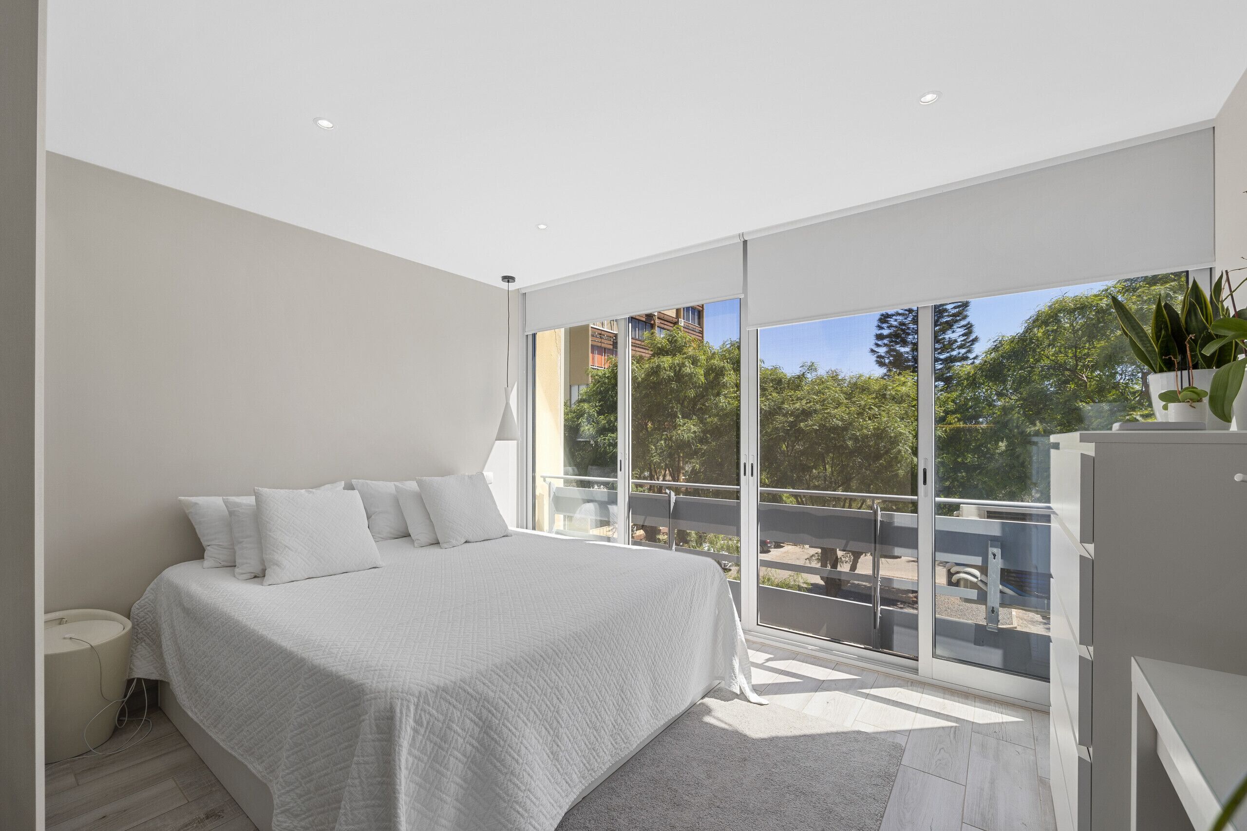 A modern bedroom with a large bed, a bedside table, and a view of a balcony with trees and buildings.