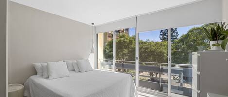 A modern bedroom with a large bed, a bedside table, and a view of a balcony with trees and buildings.