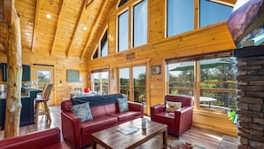Una cabaña de madera espaciosa con techo alto, ventanas grandes, un área de estar acogedora con sofás de cuero rojo, una mesa de centro de madera y una chimenea de piedra.