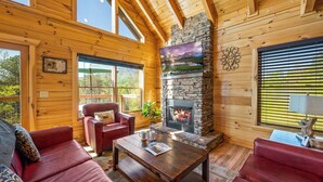 Interior acogedor de una cabaña de madera con chimenea de piedra, sofás de cuero y grandes ventanales que ofrecen vistas al exterior.
