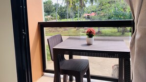 Un balcón con una mesa y sillas de mimbre, una planta en maceta y vistas a palmeras y casas.