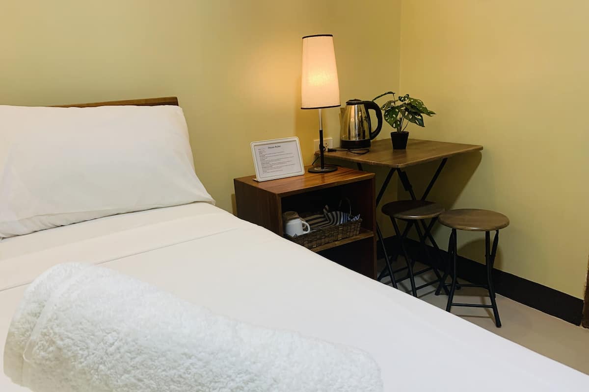 Hotellihuone, jossa on sänky, yöpöytä lampun kanssa, vedenkeitin, kasvi ja pieni jakkara.