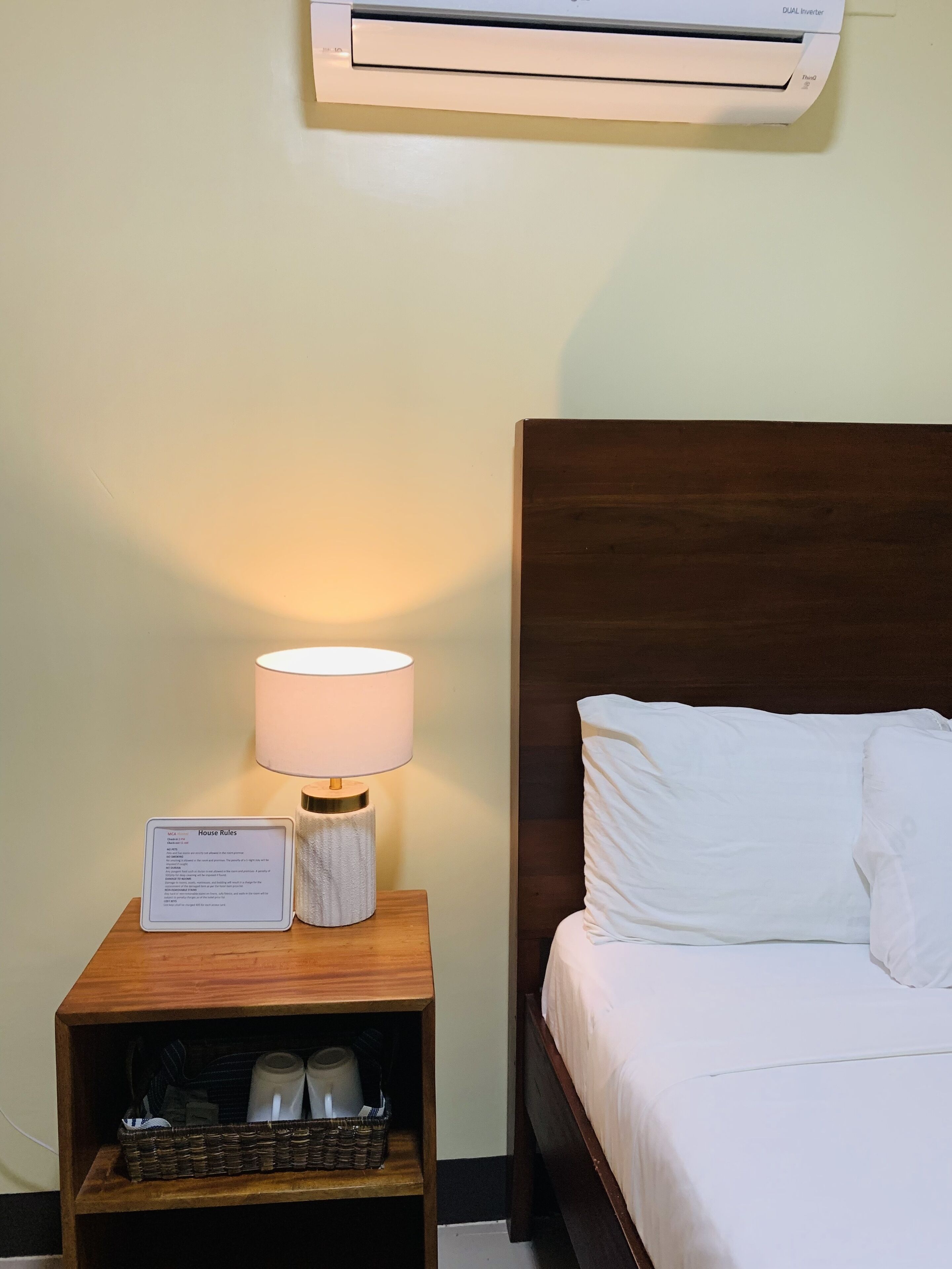 A hotel room with a bed, a nightstand with a lamp, a sign with house rules, and an air conditioner above the bed.