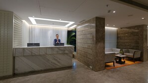 Uma área de recepção moderna com um balcão de mármore, um espaço de espera com uma planta em vaso e uma parede grande com linhas horizontais.