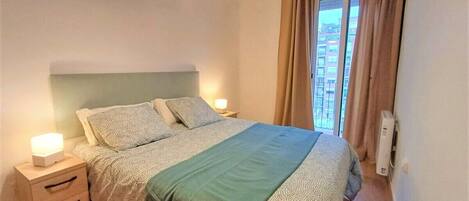 A bedroom with a bed, bedside tables, a nightstand, a window with curtains, and a wall-mounted air conditioning unit.