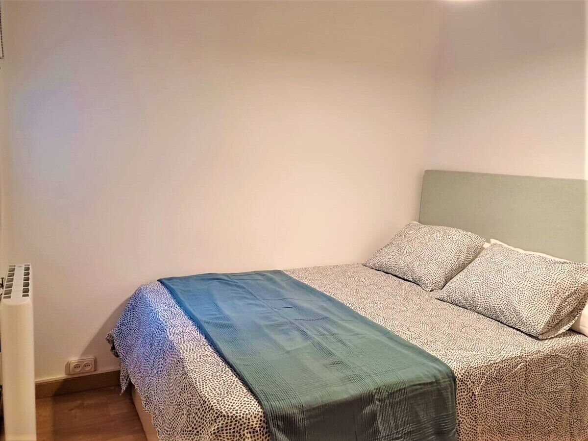 A single bed with a patterned bedspread and pillows, a green throw blanket, and a wall-mounted air conditioning unit.