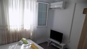 A hotel room with a bed, a television on a stand, an air conditioning unit, and a window with curtains. - VS studios Economy Double Room (Skiathos)