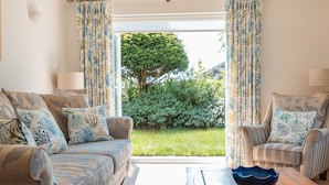 A living room with a sofa, armchair, and coffee table, a view of a garden through a glass door, and a lamp on the side table. - The Beach House - a stone's throw from Avon Beach, Friars Cliff (Christchurch)