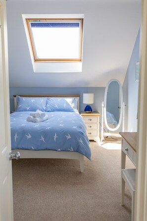 A bedroom with a skylight, a bed with a blue bedspread, a bedside table with a lamp, and a full-length mirror.