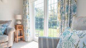 A living room with a sofa, a small wooden table with a lamp, and a view of a garden through a large window. - The Beach House - a stone's throw from Avon Beach, Friars Cliff (Christchurch)