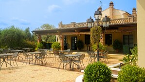 Un patio al aire libre con mesas y sillas, un edificio de ladrillo con tejado de baldosas y plantas en macetas.