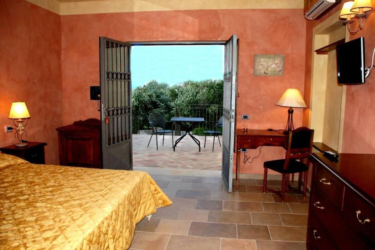 A bedroom with a bed, a desk, a chair, and an open door leading to an outdoor patio with a table and chairs.